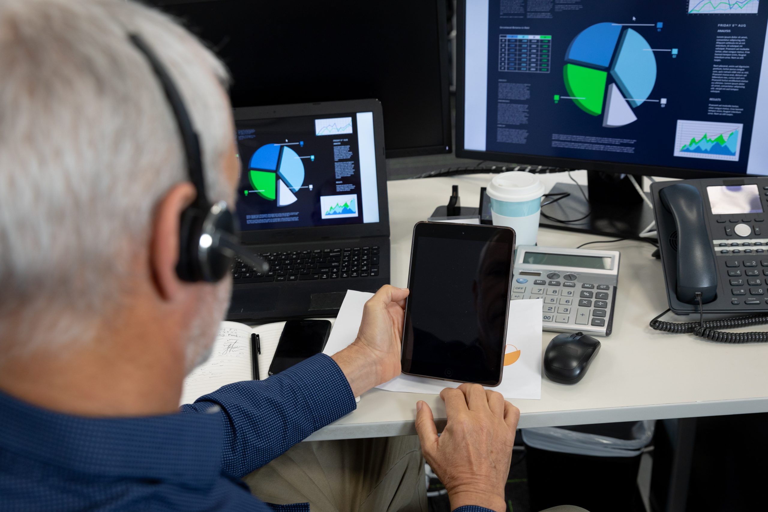 Caucasian businessman working in a modern office, sitting at desk with computer, wearing phone headset and holding tablet. Social distancing in workplace during Coronavirus Covid 19 pandemic.24/7 Monitoring